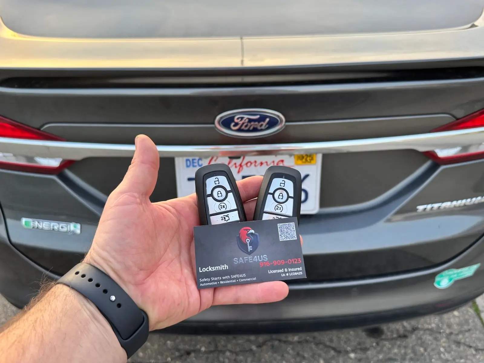 Hand holding two Ford remote key fobs and a Safe4us business card in front of a dark Ford sedan