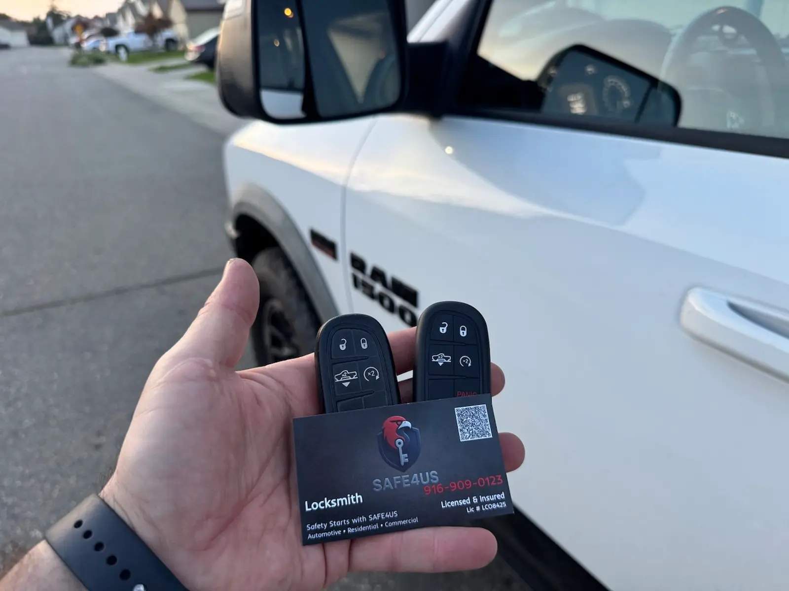 Hand holding two Ram truck key fobs and a Safe4us business card in front of a white Ram 1500